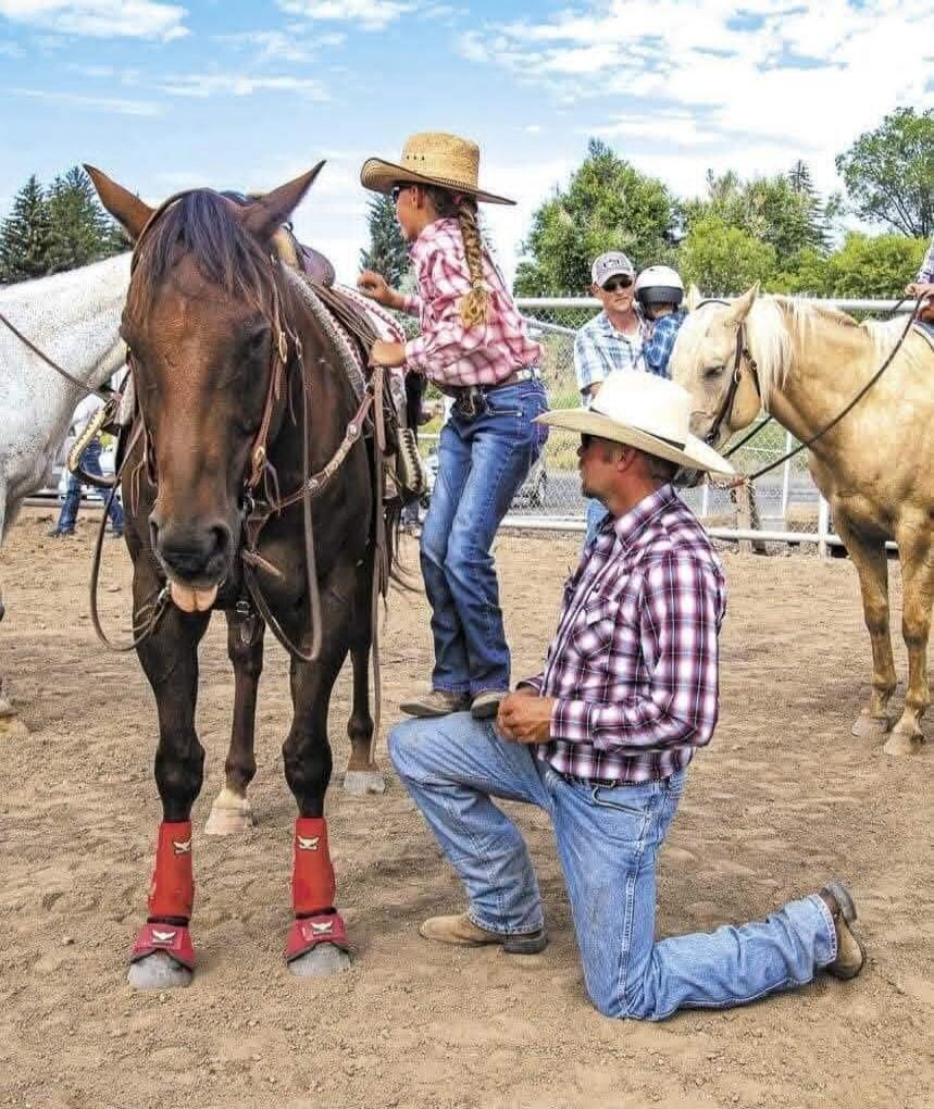 A young girl in a cowboy hat and plaid shirt is standing on a horse, while a man in a cowboy hat and plaid shirt kneels beside the horse, assisting her. The horse has its tongue sticking out in a funny way. Other people and horses are in the background at what appears to be a ranch or arena.