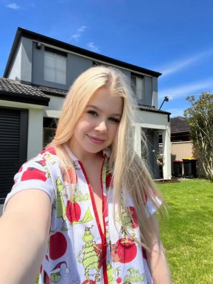 A young blonde woman takes a selfie outdoors in front of a modern house. She is wearing a white onesie with green Grinch characters and red cherry patterns. The text 'The GRING' is visible on her clothing. The sky is clear blue, and there is green grass in the foreground.