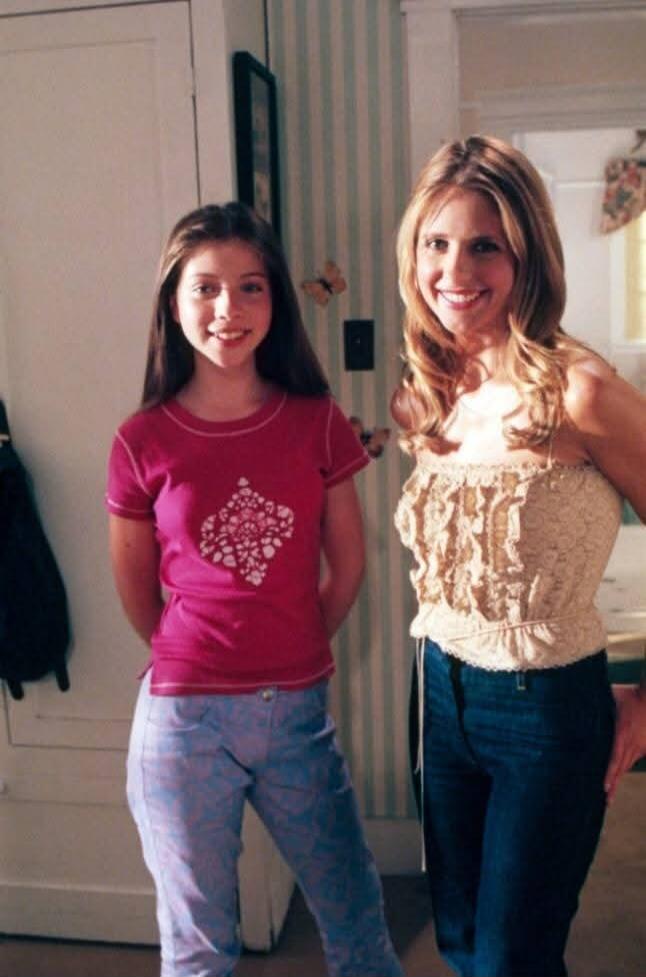 Two people posing indoors: a girl in a pink t-shirt and light blue jeans standing next to a woman wearing a beige lacy strapless top and dark blue jeans.