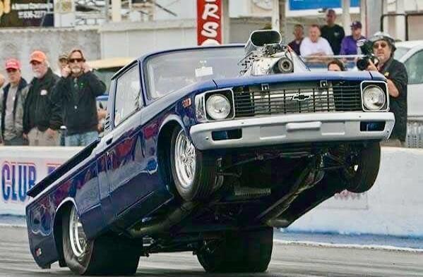 A blue drag racing pickup truck performs a wheelie on a drag strip. Spectators are watching in the background. Text visible: CLUB, RS.