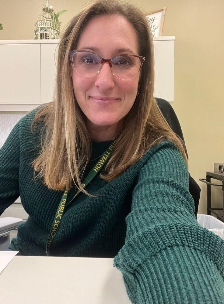 A woman wearing glasses and a green sweater, smiling at the camera. She has a lanyard around her neck that reads 'HOWELL PUBLIC SCHOOLS'.