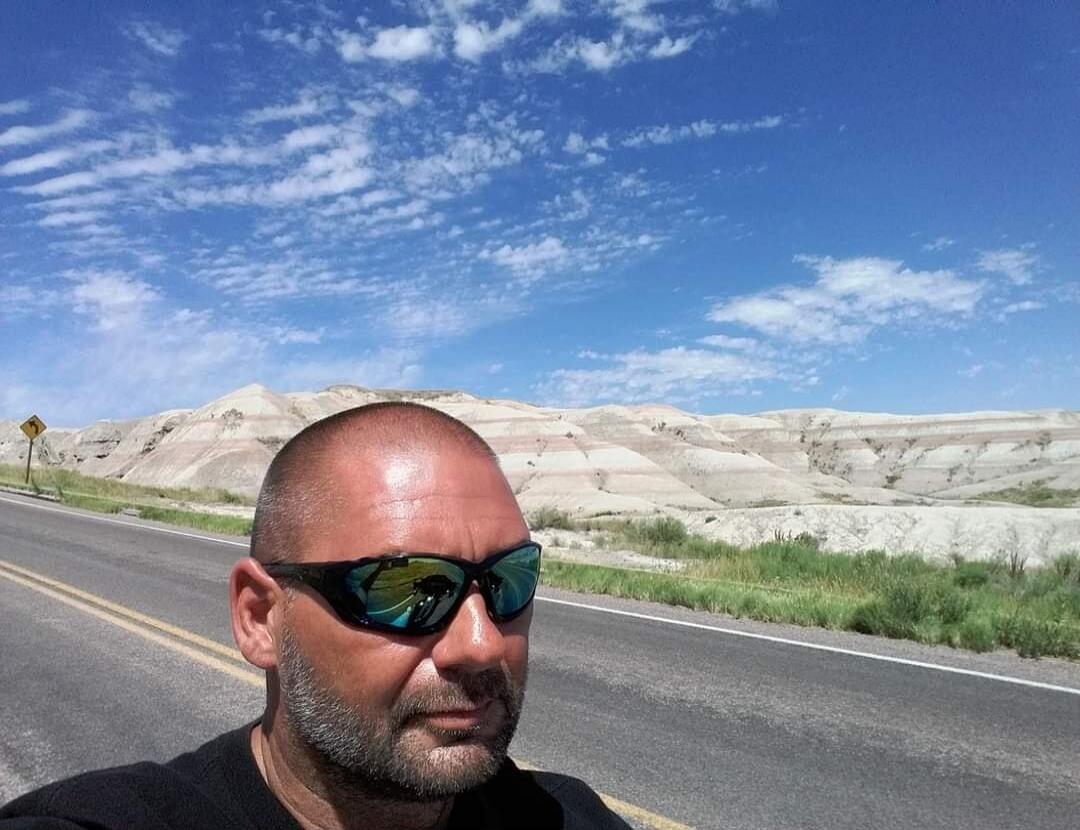 A man in sunglasses is taking a selfie on a road with a mountainous landscape in the background.