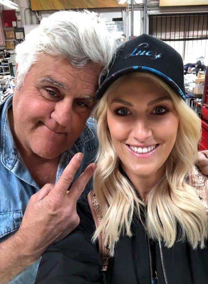 Jay Leno and a blonde woman taking a selfie. The woman is wearing a black cap with 'Lucy' written on it. Jay Leno is making a peace sign.