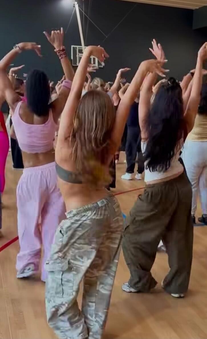 A group of people, mostly women, are seen from behind in a dance or fitness class, with their arms raised in a synchronized movement.