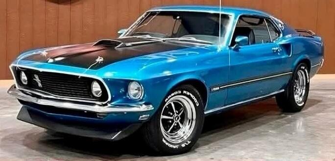 A classic blue Ford Mustang, likely a 1969 or 1970 model, is parked indoors. The car features a black hood with a scoop and chrome trim. The tires have 'COOPER COBRA RADIAL G/T' written on their sidewalls.