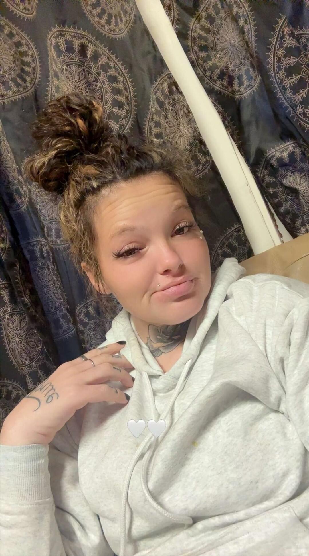 A woman with a top bun hairstyle, wearing a gray hoodie, sitting on a couch.