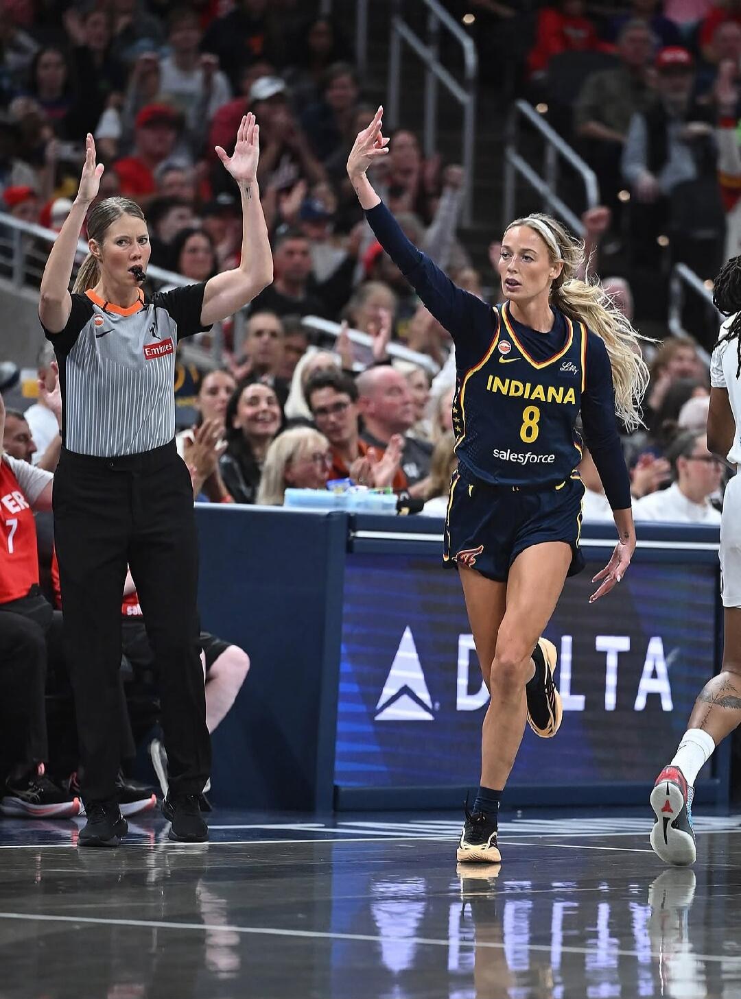 A female referee with an Emirates logo on her shirt is making a hand signal on a basketball court. A female basketball player wearing an Indiana Fever jersey with number 8, sponsored by Salesforce and Lilly, is also making a hand gesture. A Delta logo is visible in the background. Spectators are in the stands.