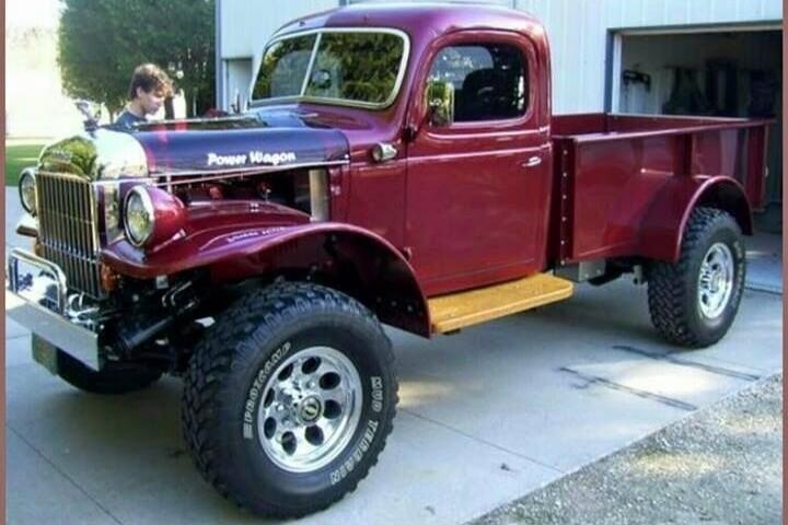 Power Wagon. Mud Terrain. A classic red Dodge Power Wagon pickup truck, heavily modified with large off-road tires and a lifted suspension, is parked on a concrete driveway in front of a garage. A person is visible near the front of the truck.