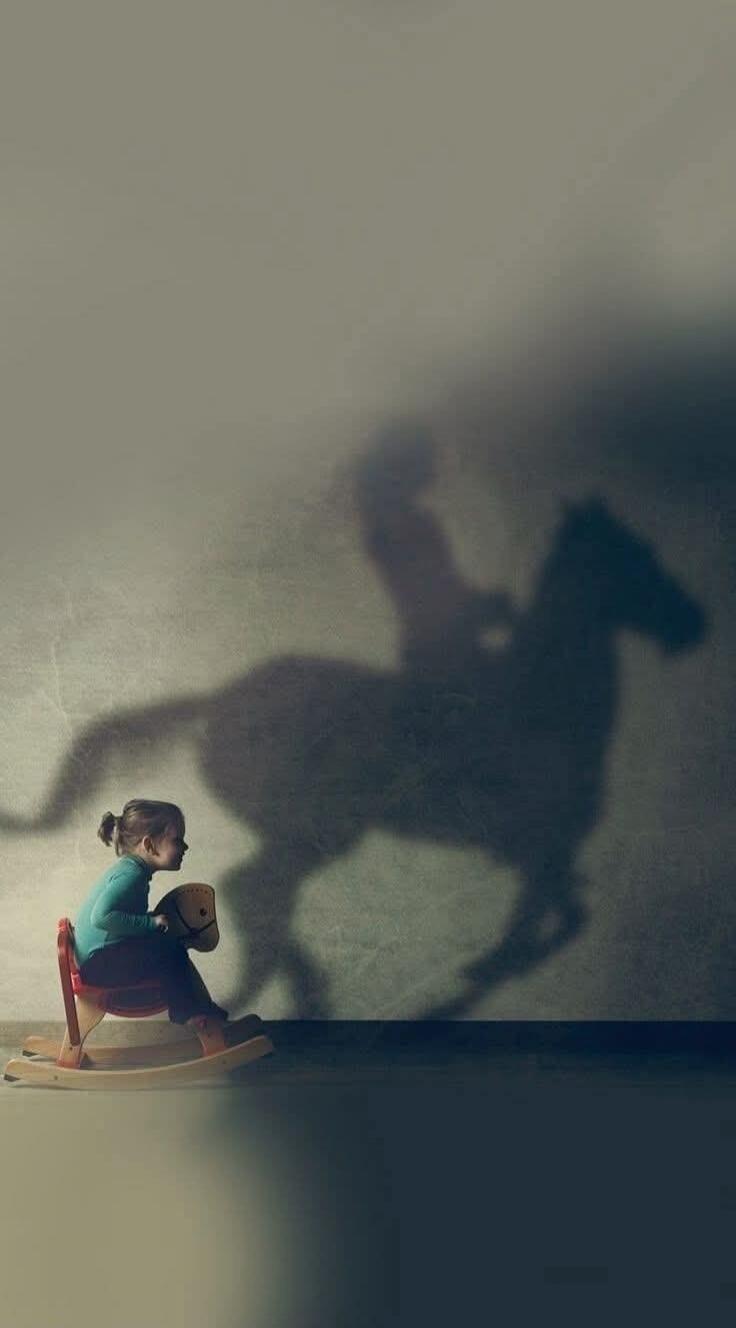 A child on a rocking horse with a large shadow of a rider on a wall.