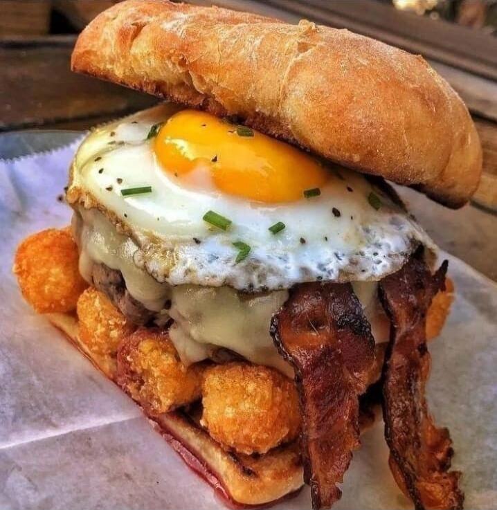 A loaded burger topped with a sunny-side-up egg and chives. The patty is layered with melted cheese, crispy bacon, and appears to be surrounded by tater tots inside the bun, plated on parchment.