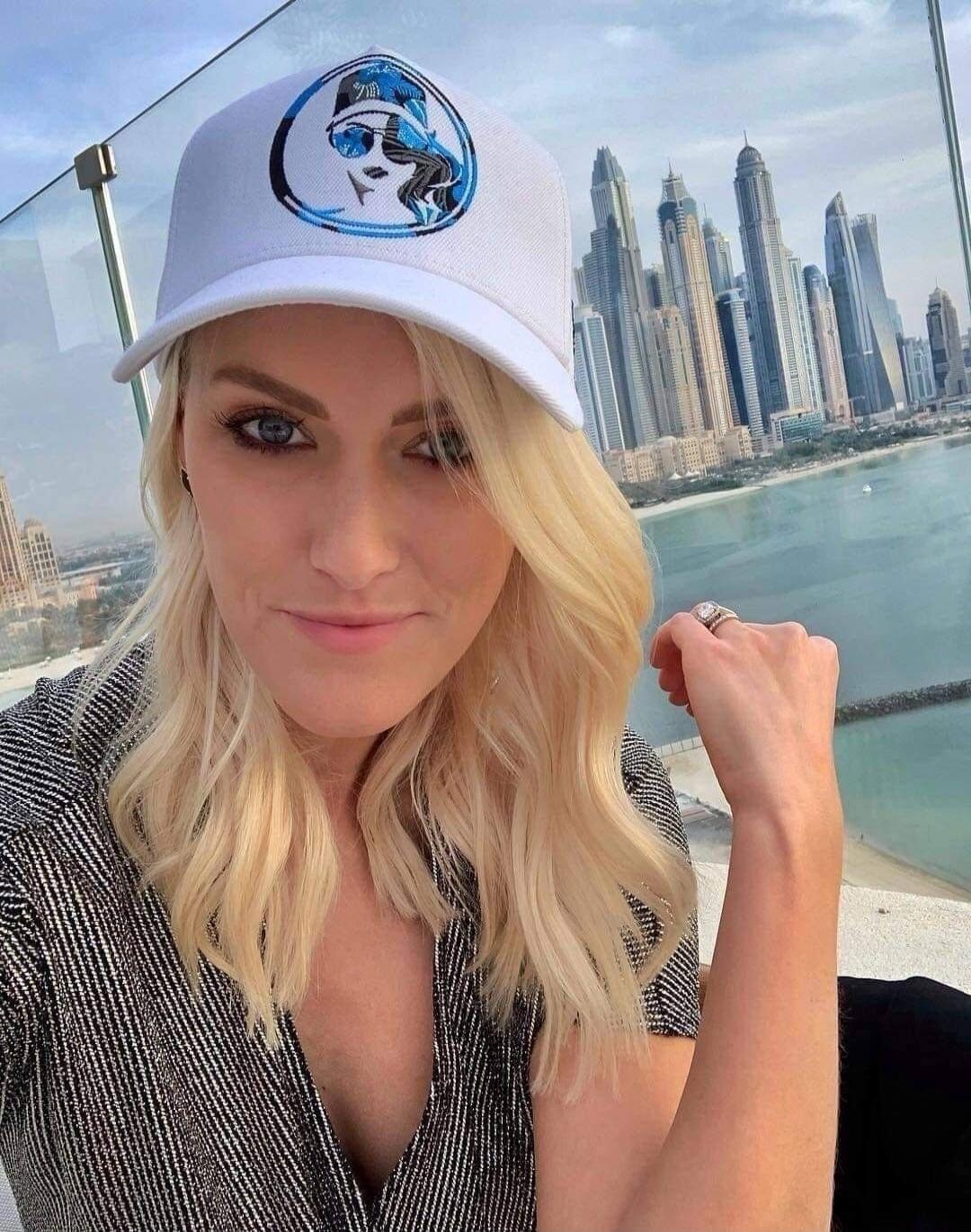 A blonde woman takes a selfie with a city skyline and water in the background. She is wearing a white cap with a blue and black logo and a striped top.