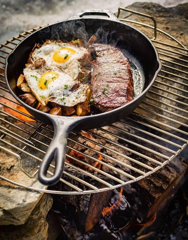 Two fried eggs and a steak cooking in a skillet over an open flame.