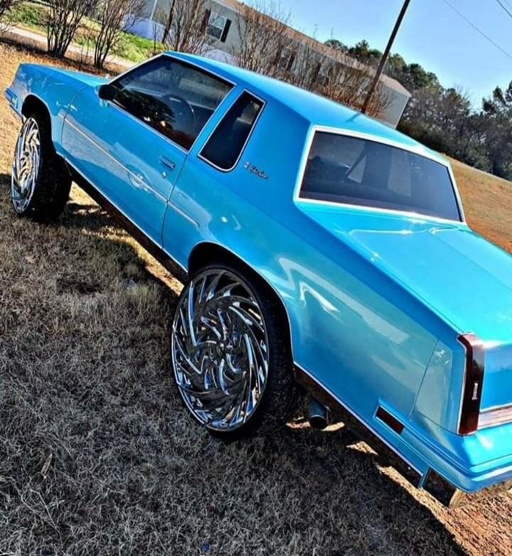 The image shows a bright blue classic car with large chrome rims parked on a grassy area. The car appears to be a coupe with a distinctive body style. The wheels are highly polished and have a complex, swirling design. The background shows some trees and houses, suggesting a suburban or rural setting. The sky is clear and blue.