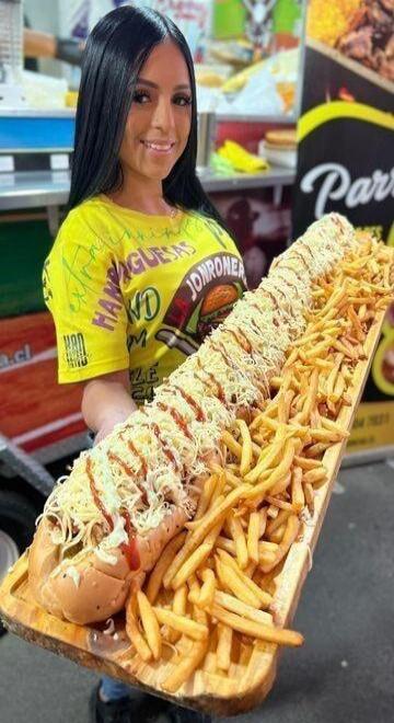 A smiling woman holding a giant hot dog with cheese and sauce, served with french fries. Text on her shirt includes 'HAMBURGUESAS' and 'JONRONER'. A sign in the background shows 'Parri'.