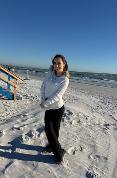 A young woman stands on a sandy beach, smiling at the camera. She is wearing a white sweater with a fur-trimmed hood and black pants. The ocean and a clear blue sky are visible in the background.