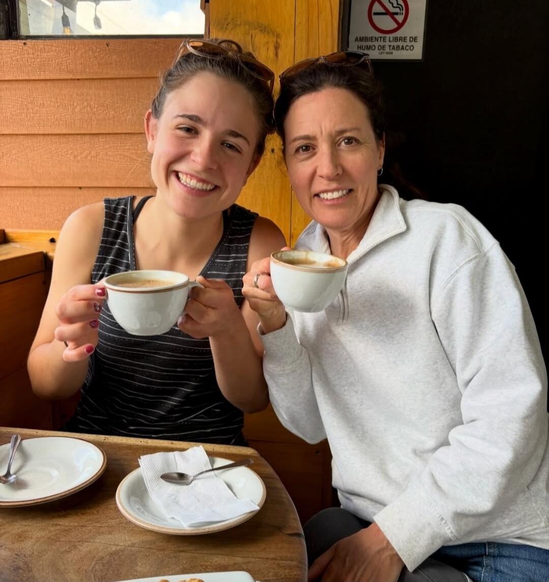 Two smiling women are sitting at a wooden table, holding coffee cups. In the background, there is a sign with a no-smoking symbol and the text 'AMBIENTE LIBRE DE HUMO DE TABACO', which means 'Tobacco smoke-free environment'.