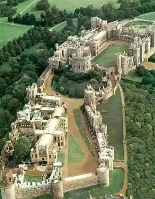 Aerial view of a large medieval-style castle complex with towers and a moat, surrounded by green lawns and dense trees.