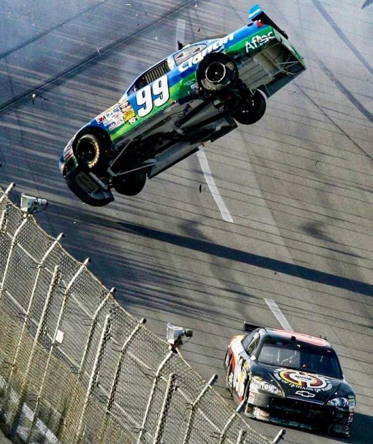 A race car, number 99, is airborne and upside down on a race track, possibly after a crash. Another car is visible on the track below. Debris is flying around the car. Visible text includes '99', 'Cran', 'Aflac', '3M', 'ISC', and 'Chevrolet'.
