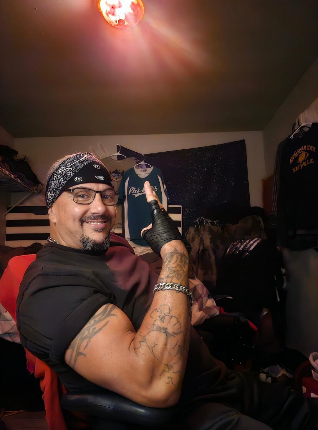 A man with a bandana, glasses, and tattoos is smiling and pointing upwards. In the background, there are clothes hanging, including a shirt with 'Phila' and another with 'APPALACHIAN STATE FOOTBALL' visible.