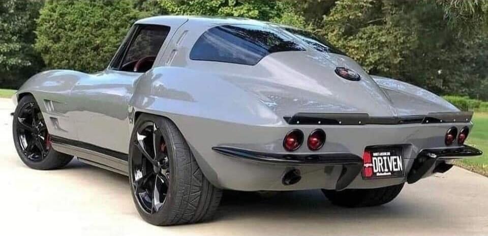 A grey classic Chevrolet Corvette C2 Sting Ray with custom black wheels and a license plate that reads 'DRIVEN'. The car is parked outdoors on a paved surface with green trees in the background.