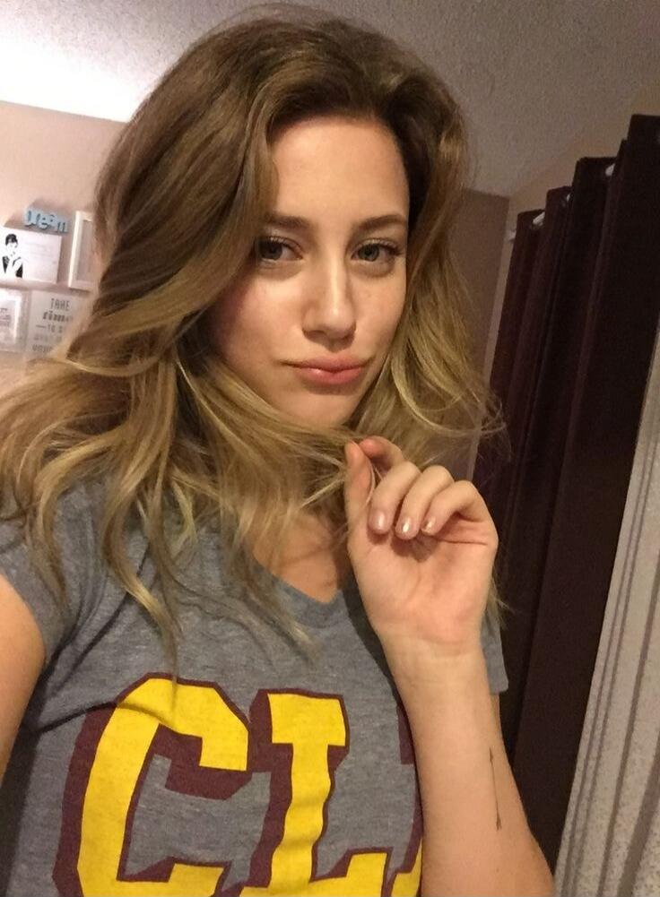 A young woman with long blonde hair takes a selfie, wearing a grey t-shirt with yellow and maroon letters. She is looking at the camera and gently touching her hair. A small arrow tattoo is visible on her wrist. In the background, there are framed items on a shelf with text like 'dream' and 'TAKE TIME'.