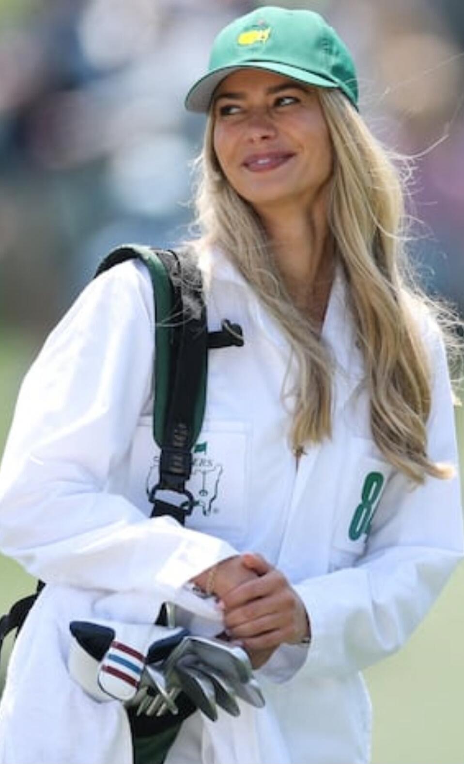 A woman wearing a green Masters golf cap and a white caddie uniform with the number 8 on the sleeve, holding golf clubs.