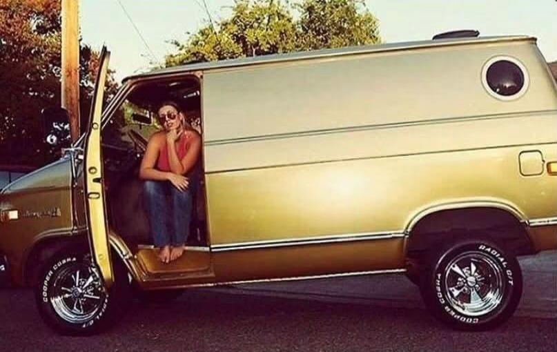 A woman is sitting in the open door of a vintage gold and brown van. She is wearing a red top, jeans, and sunglasses, with bare feet. The van has custom wheels with 'COOPER COBRA RADIAL G/T' written on the tires.