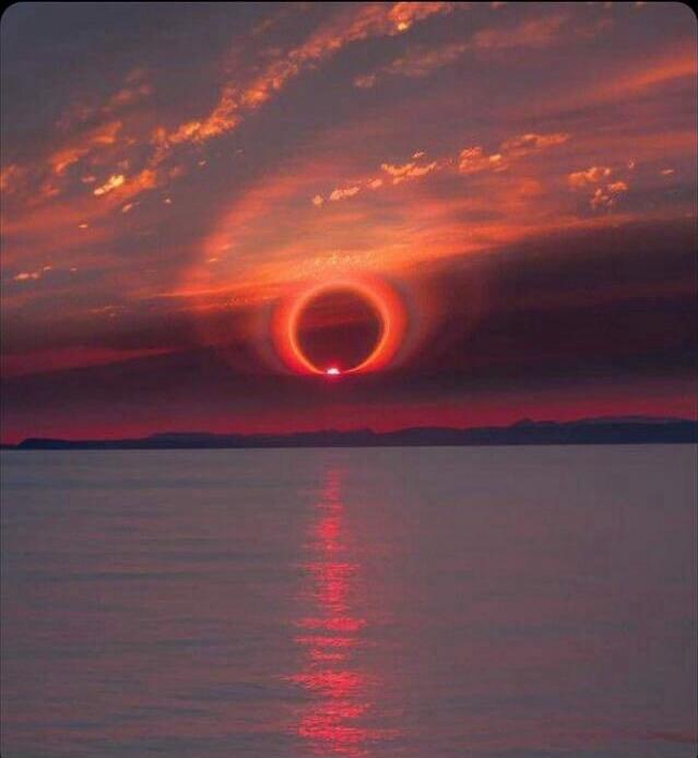 A dramatic sunset over a calm lake with a striking ring around the sun, resembling a solar eclipse.