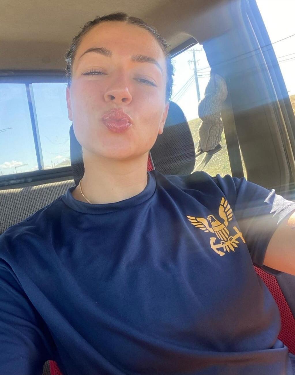 A young woman in a car taking a selfie, making a kissing face. She is wearing a dark blue t-shirt with a golden emblem.