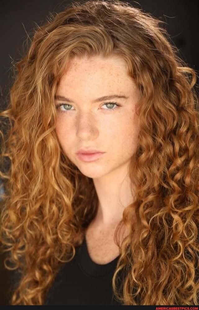 A young woman with long red curly hair and freckles, wearing a black shirt, looking at the camera.