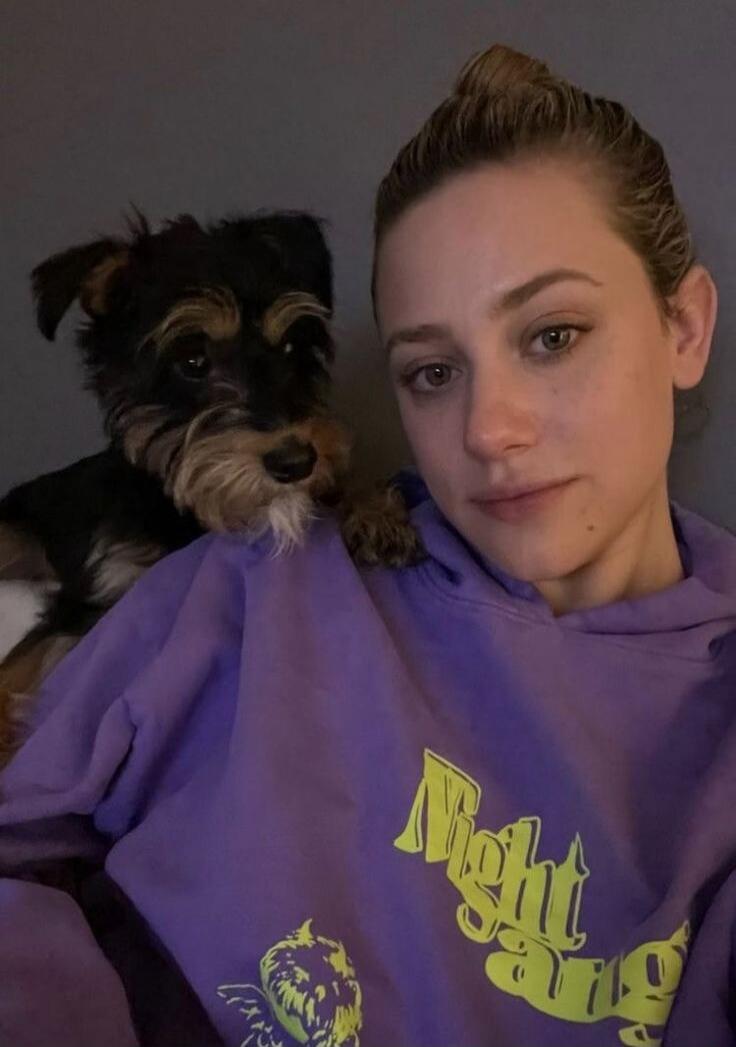 A woman taking a selfie with a small dog on her shoulder. She is wearing a purple hoodie with the text 'Night Gang'.