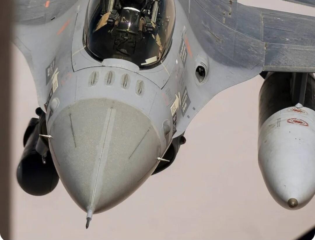 A close-up, low-angle view of the front and underside of a grey fighter jet, possibly an F-16, in flight. The cockpit canopy is visible, reflecting light. External stores, likely fuel tanks or missiles, are attached to the wings. Various sensors and warning labels are visible on the aircraft's surface.