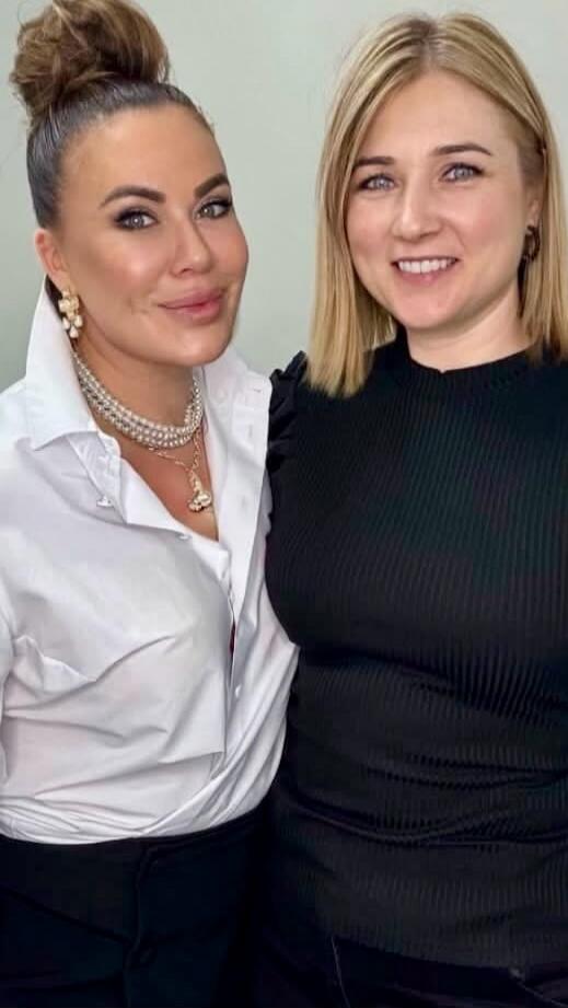 Two women smiling, one with dark hair in a bun wearing a white shirt and pearl necklaces, the other with blonde hair wearing a black ribbed top.