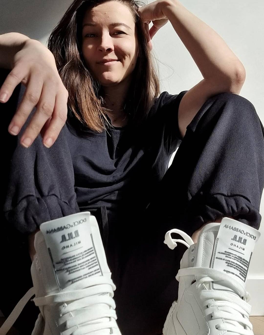 A person sitting with knees up, wearing dark clothing and oversized white sneakers with visible labels on the tongue.