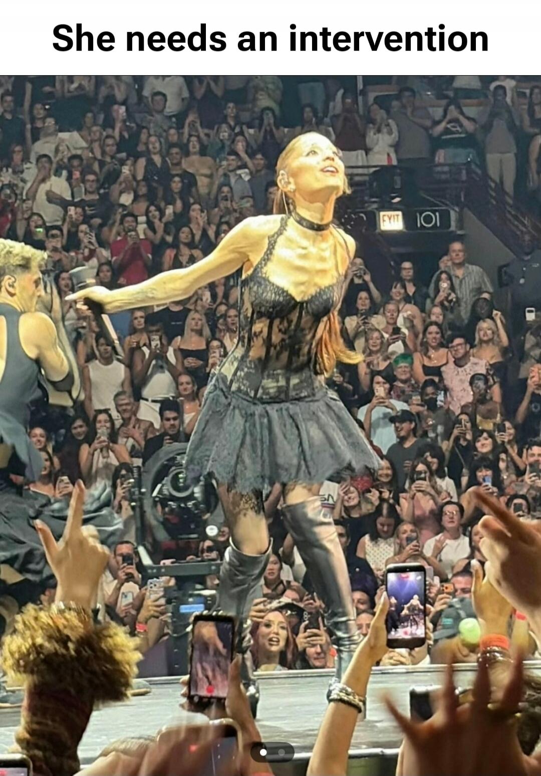 She needs an intervention. A very thin woman, possibly a celebrity like Madonna, is performing on stage in a black lace outfit and boots, looking up with her arms outstretched. A large crowd is visible in the background, many holding up phones to record the performance.
