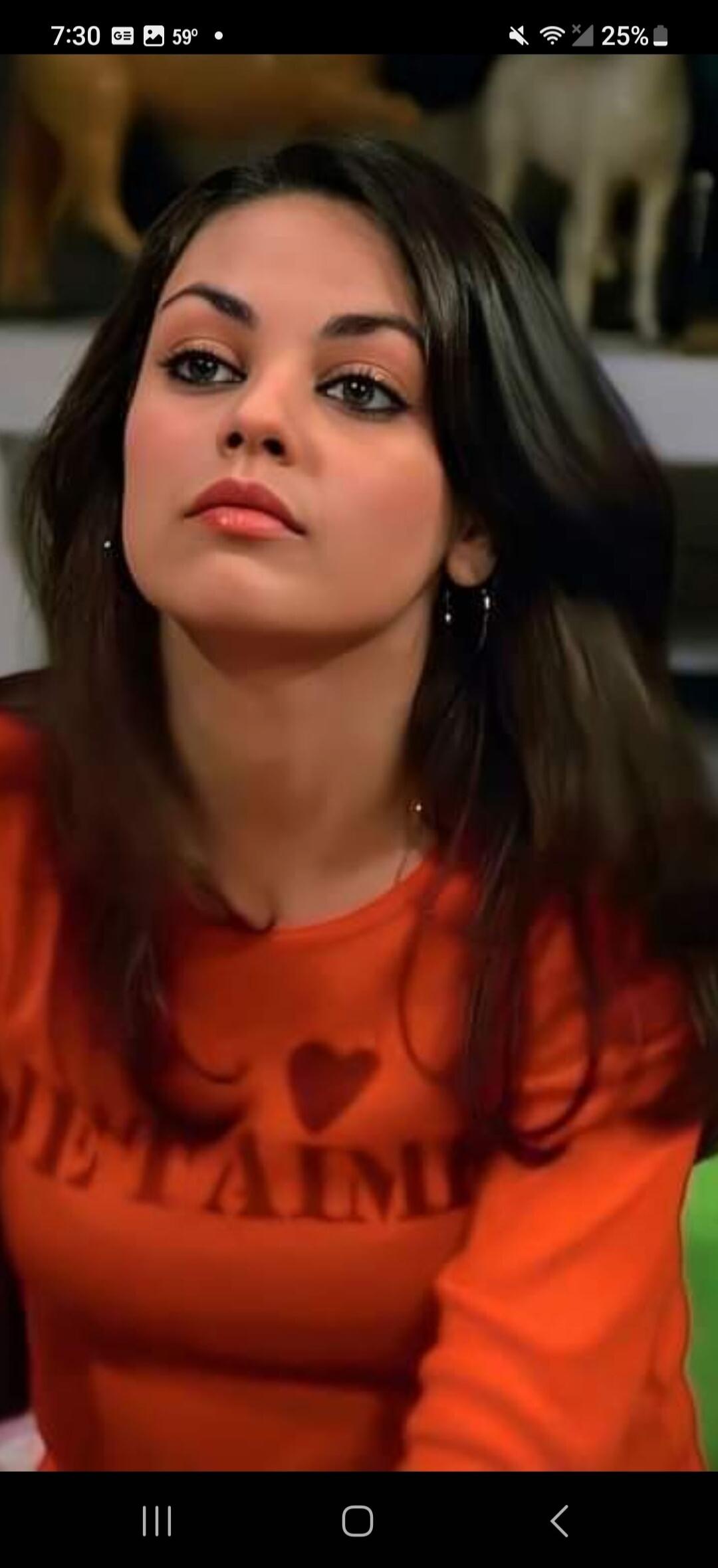A woman, resembling Mila Kunis, with dark hair and prominent eyes, wearing an orange long-sleeved shirt with a heart graphic. The shirt also has the word 'ETAM' partially visible.
