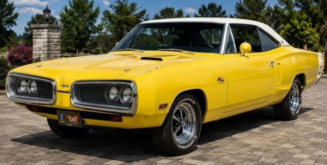 A classic yellow muscle car with a white roof is parked on a paved surface outdoors.