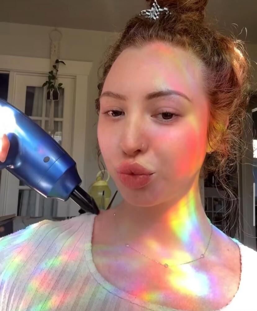 A woman with a blue hair dryer near her face, colorful rainbow light reflections across her skin.