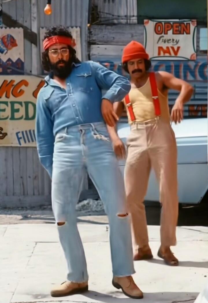 Two men, resembling Cheech and Chong, are dancing or posing outdoors. The man on the left wears a red bandana, glasses, a beard, a denim shirt, and ripped light blue jeans. The man on the right wears a red beanie, a mustache, a yellow tank top with red suspenders, and light brown pants. In the background, signs read 'OPEN EVERY MV' and partially vi