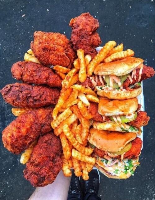 A large tray of fried chicken pieces, crinkle-cut fries, and stacked cheeseburger sandwiches. The items are piled high and displayed as a feast.