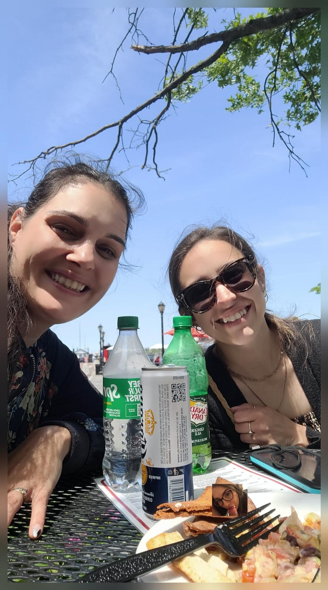 Two women are smiling at the camera, sitting at an outdoor table. On the table are bottles of Sprite, a can of Corona beer, and a can of Canada Dry. There is also a plate with food. The sky is blue with tree branches visible in the background.