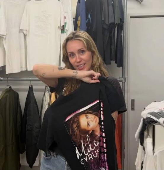 Miley Cyrus is holding a black t-shirt with her own image and name 'Miley Cyrus' printed on it. She is smiling and looking at the camera in a room with clothing racks.