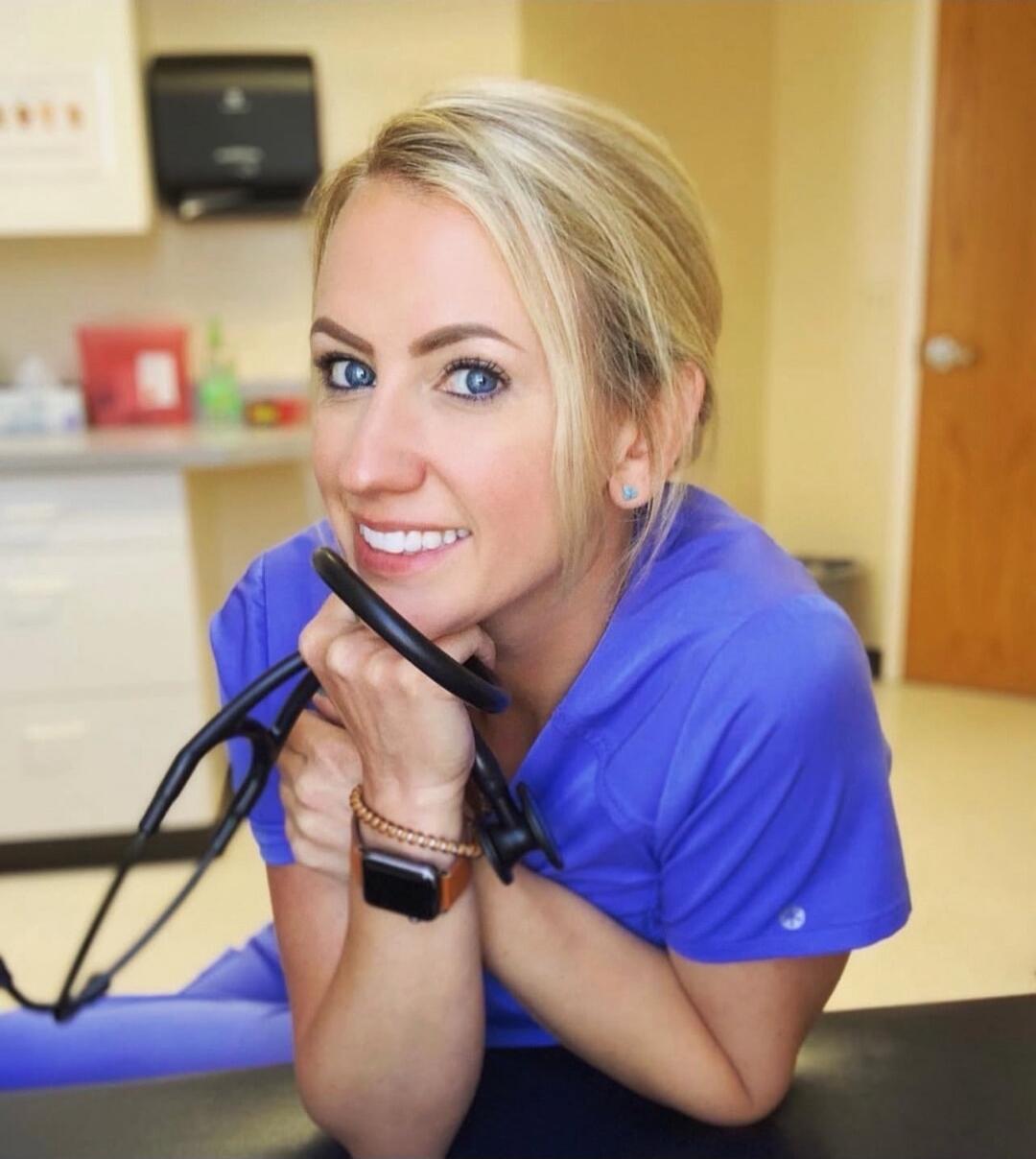A woman in blue scrubs smiling while holding a stethoscope in a clinical setting.