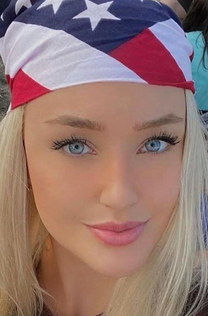A close-up portrait of a blonde woman wearing a bandana with the American flag pattern. She has blue eyes, long lashes, and pink lips.