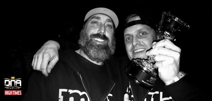 Two men are smiling, one holding a trophy from the High Times Amsterdam 30th Anniversary Seed Competition. The logo 'DNA GENETICS HIGH TIMES' is visible in the bottom left corner.