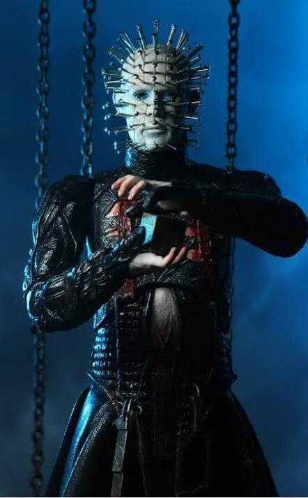 Figure wearing a spiked mask with pins all over, dressed in dark gothic attire, standing among chains.