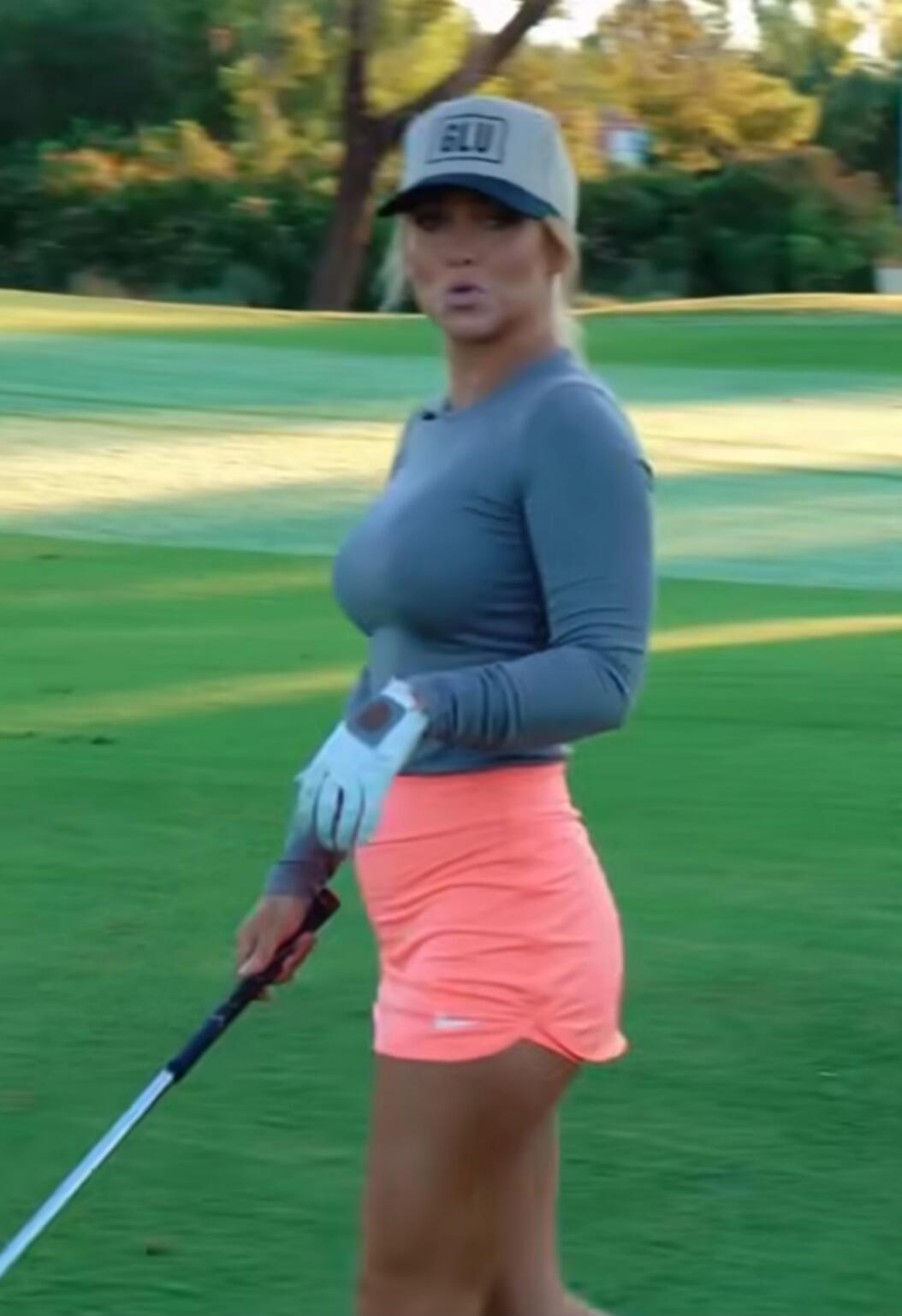 Image shows a female golfer on a golf course. She is wearing a gray long-sleeve top, bright pink/orange shorts, a light-colored cap, and white golf glove, holding a golf club.