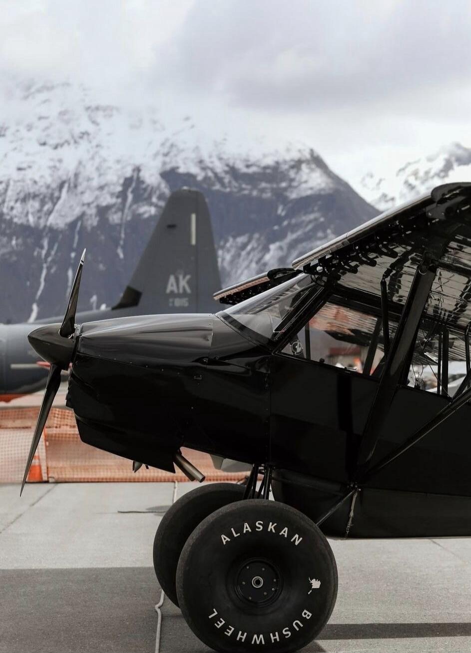 A black bush plane with 'ALASKAN BUSHWHEEL' written on its large tires is parked on an airfield. In the background, a military transport aircraft with 'AK' on its tail is visible, set against a backdrop of snow-capped mountains under a cloudy sky.