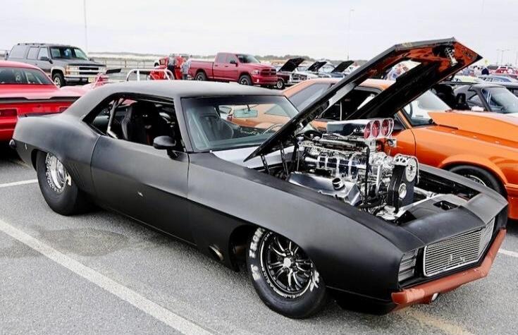 Matte black muscle car with the hood and front deck opened to reveal a large chrome engine display. Custom wheels, low stance, and a sleek, aggressive look at a car show.