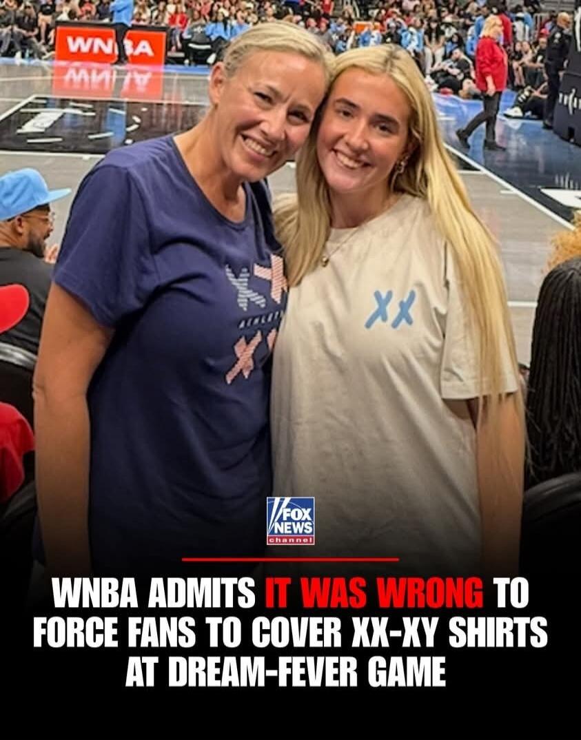WNBA ADMITS IT WAS WRONG TO FORCE FANS TO COVER XX-XY SHIRTS AT DREAM-FEVER GAME. Two women are smiling at a WNBA game. One woman wears a dark blue shirt with an 'XX' pattern, and the other wears a light-colored shirt with an 'XY' pattern. The WNBA logo is visible on the court in the background.